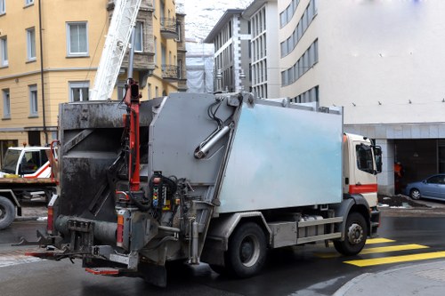 A large commercial waste collection truck is parked on a city street, positioned next to a zebra crossing with yellow lines. The truck has a grey metal body with visible wear and grime, particularly around the rear loading area where hydraulic components and mechanisms are exposed. The rear section includes a black loading hatch that appears open or accessible for waste loading, with hydraulic arms and controls visible at the sides. The vehicle is situated in front of a row of multi-storey buildings with varied facades, including a yellow building to the left and modern glass structures to the right, suggesting an urban environment such as Turnham Green. Part of a red ladder is mounted on the truck’s side, indicating it may be equipped for various rubbish collection tasks. The street surface appears wet, reflecting the overcast sky, which contributes to the muted lighting. Nearby parked cars are visible, consistent with typical urban waste collection scenes. This image illustrates the type of commercial waste removal service provided by Commercial Waste Turnham Green in the area, focused on urban rubbish collection and disposal.