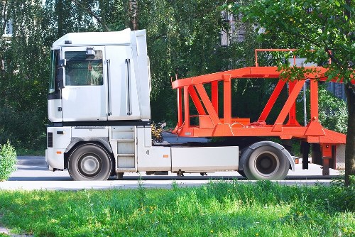 A white and grey commercial lorry with an orange open flatbed trailer is parked on a paved surface adjacent to a grassy area with small yellow flowers. The trailer, designed for waste collection, features a metal railing around its perimeter and is partially covered with green tree branches extending into the frame. The background includes leafy green trees and a hint of a building, indicating a suburban or urban environment, possibly near Turnham Green. The vehicle is clean, and the scene is illuminated by daylight, emphasizing its functionality for rubbish removal services in the local area served by Commercial Waste Turnham Green.