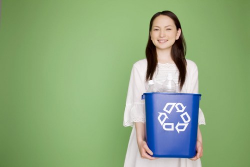 A woman standing outdoors against a plain green background, holding a blue plastic recycling bin with a white recycling symbol printed on the front. She is dressed in a white, short-sleeved dress and has long, dark hair. The bin is empty and held at waist level, with her hands gripping the sides. The setting appears to be in a residential or commercial area with natural lighting, emphasizing the importance of waste separation and responsible rubbish disposal, consistent with services provided by Commercial Waste Turnham Green for rubbish removal within the local area of Turnham Green. The image conveys a professional attitude towards recycling and rubbish management, aligned with environmentally conscious waste practices in the local community.