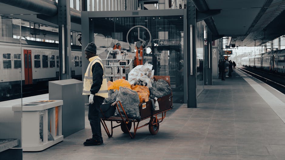 A waste collection worker wearing a yellow safety vest and dark trousers is positioned in front of a large red refuse collection vehicle, which is parked on the side of a paved road. The rear section