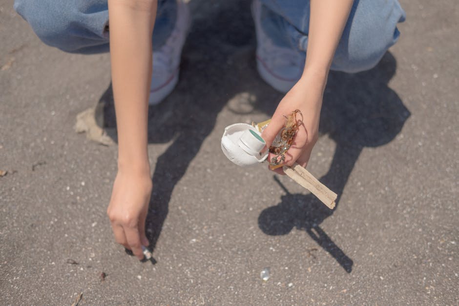 A person crouching on a paved outdoor surface, seen from the knees up, is holding a small pile of debris including a white ceramic or plaster piece, possibly part of a broken object, with visible crac